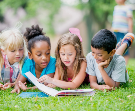 four children of different ethnicities sitting on the grass reading children's books together